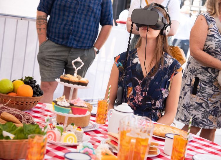 A white woman wearing a VR headset sits at a picnic table. There are cakes and glasses of orange juice on the table.